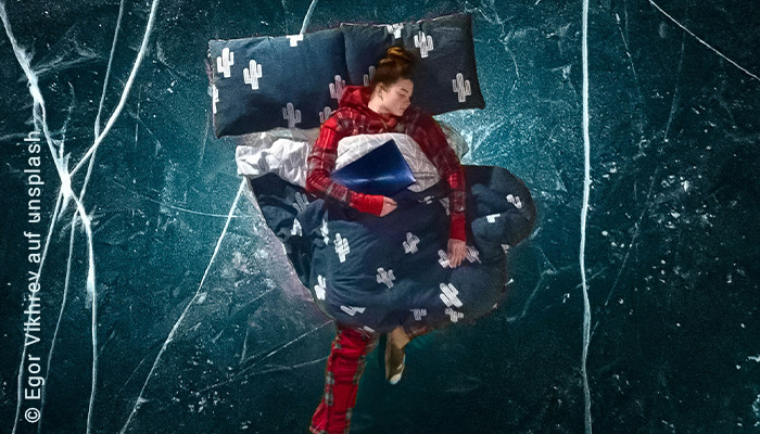 Eine Frau liegt im karierten Schlafanzug in Bettwäsche mit Kakteen-Muster in schwarz-weiß. Das Bild ist freigestellt vor einem wie schwarzer Granit anmutendem Hintergrund.