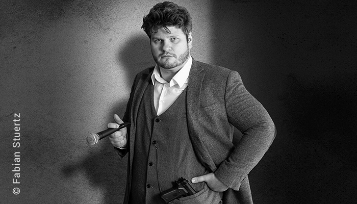Portrait des Comedian Jan Philipp Zymny in Gangsterpose: Bekleidet mit einem Dreiteiler hält er das Mikrofon wie eine Zigarre zwischen Fingern und in der Westentasche steckt ein Revolver an einer Uhrenkette